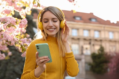 Photo of Young woman with smartphone and headphones listening to music outdoors on sunny day, space for text