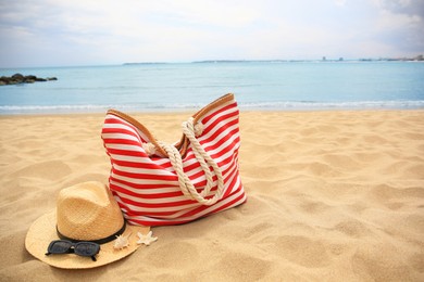 Photo of Stylish striped bag with straw hat, sunglasses, seashell and starfish on sandy beach near sea. Space for text