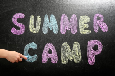 Photo of Child writing SUMMER CAMP with colorful chalk on blackboard, closeup