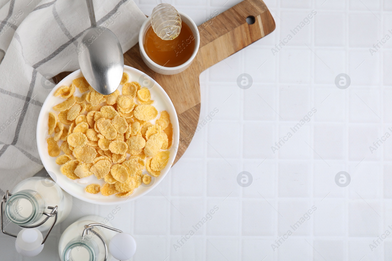 Photo of Breakfast cereal. Tasty corn flakes, milk, honey and spoon on white tiled table, top view. Space for text