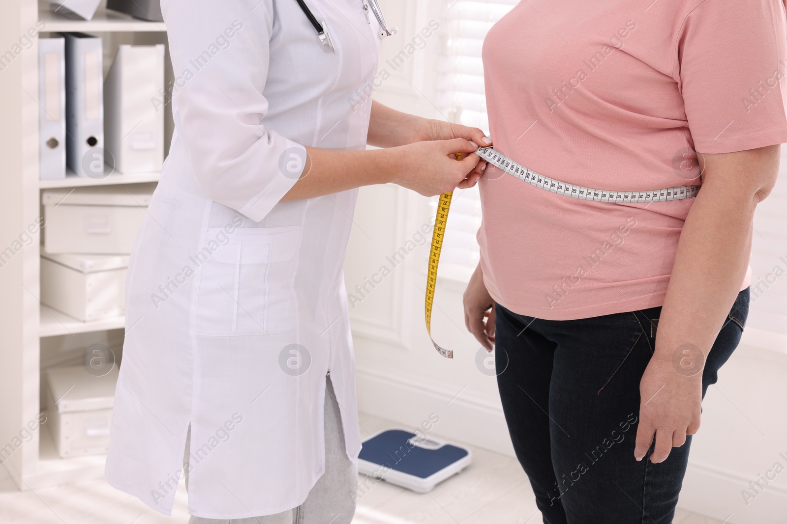 Photo of Nutritionist measuring overweight woman's waist with tape in clinic, closeup