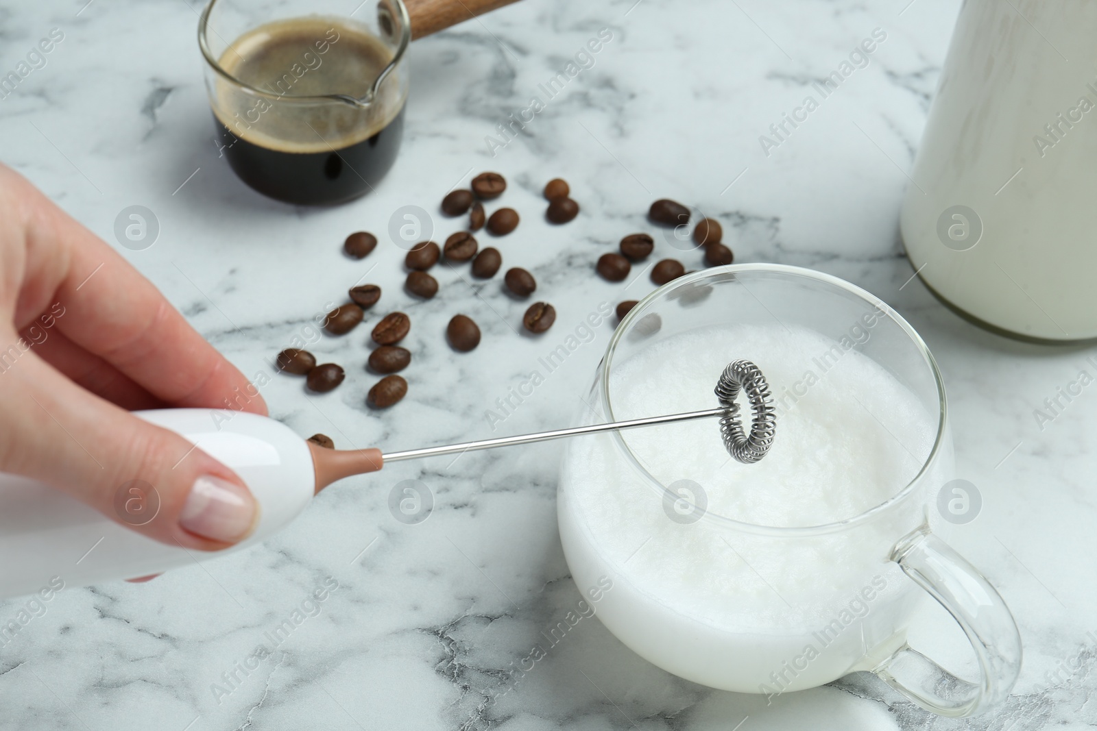 Photo of Woman whisking milk in cup with mini mixer (milk frother) at white marble table, closeup