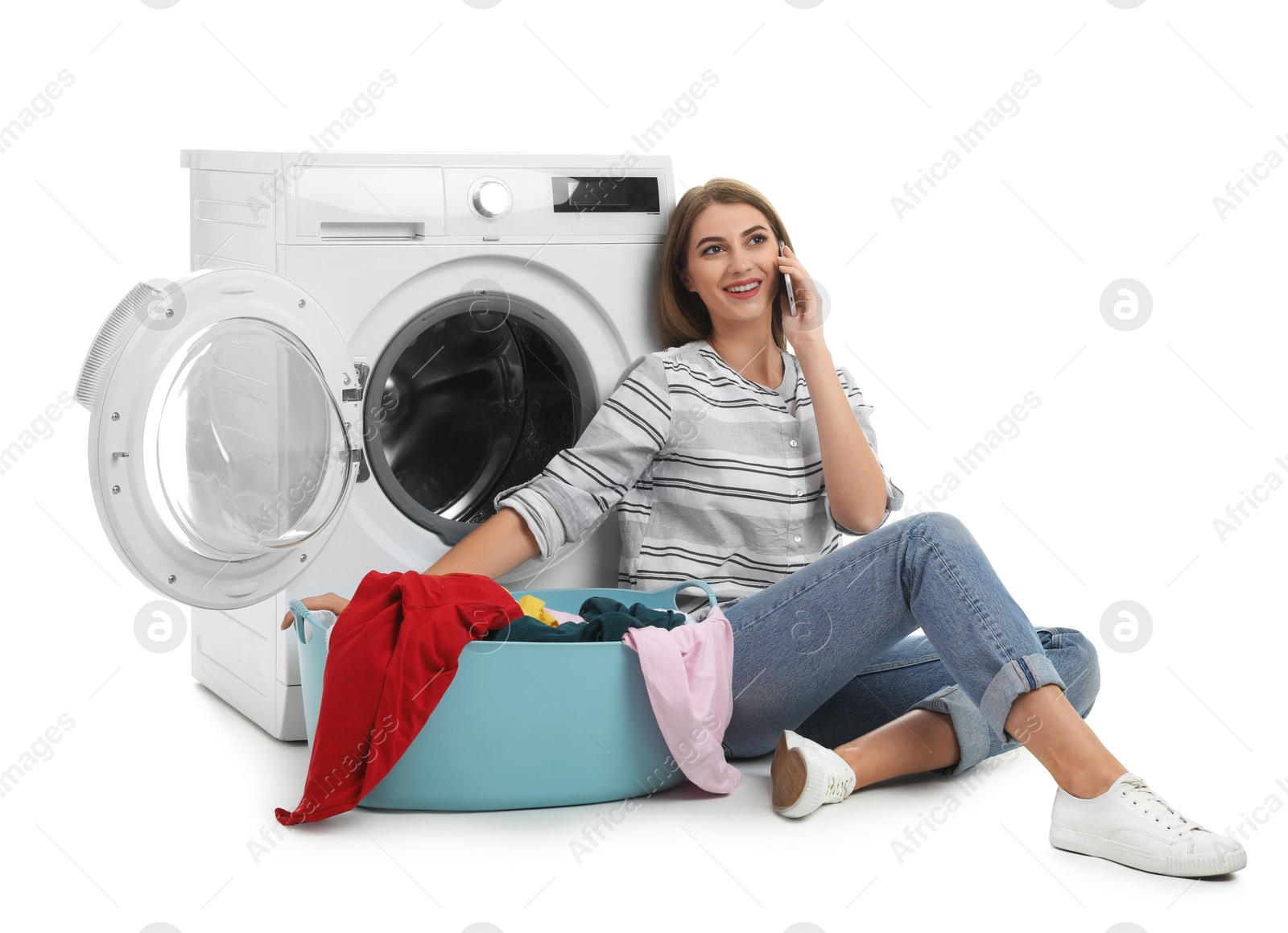 Photo of Young woman with basket of laundry sitting near washing machine and talking on phone against white background