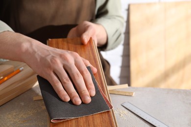 Photo of Man polishing wooden plank with sandpaper at grey table indoors, closeup