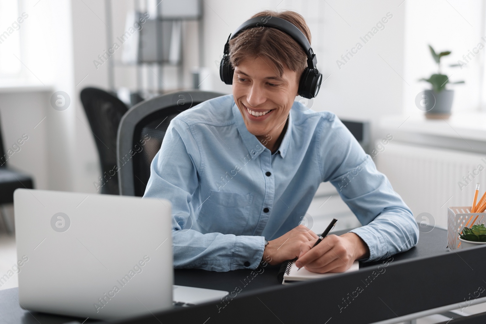 Photo of Man in headphones taking notes during webinar at table in office