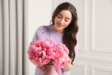 Photo of Beautiful young woman with bouquet of pink peonies in vase indoors, selective focus