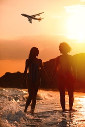 Couple walking on beach and airplane flying in sky at sunset. Summer vacation