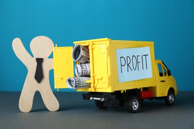 Photo of Economic profit. Wooden figure and toy truck with banknotes on grey table against light blue background