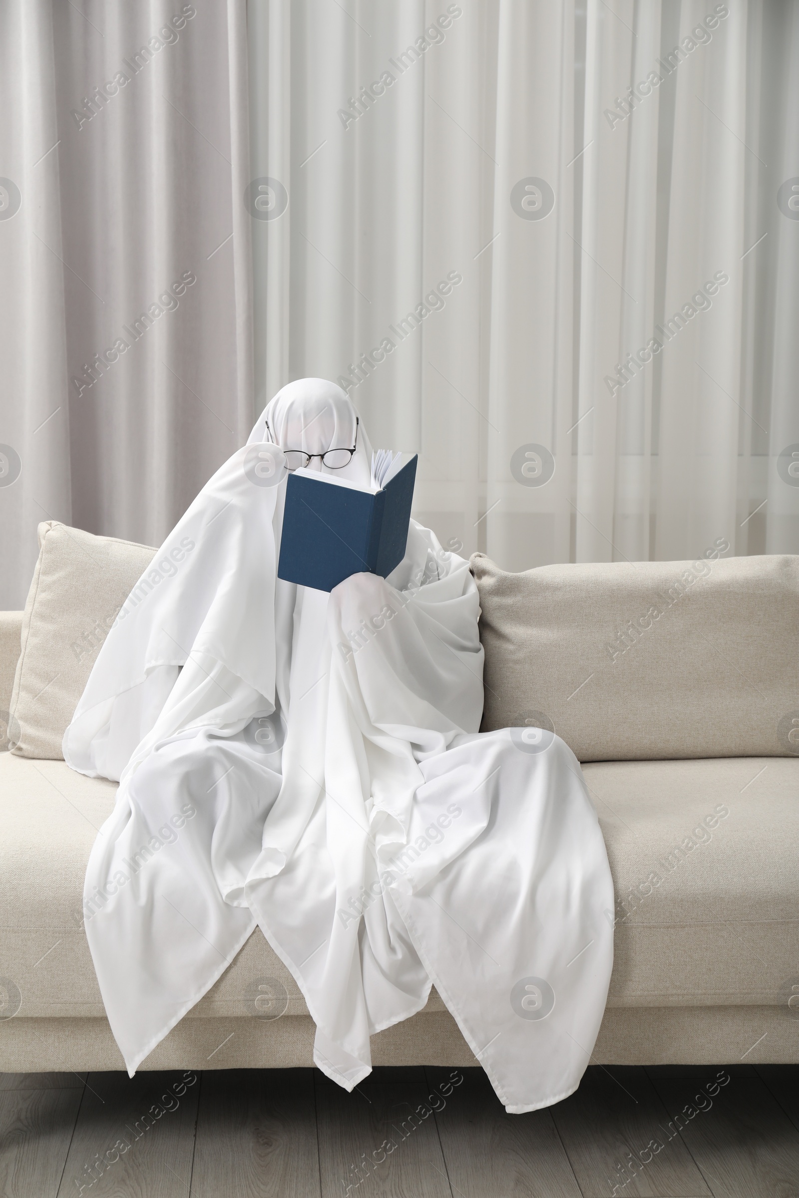 Photo of Creepy ghost. Person covered with white sheet reading book on sofa at home