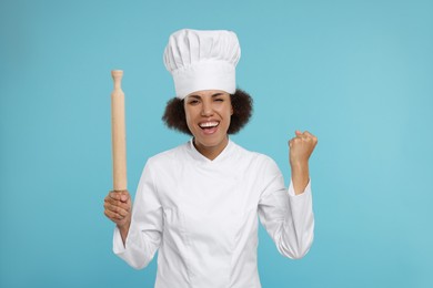 Photo of Emotional female chef in uniform holding rolling pin on light blue background
