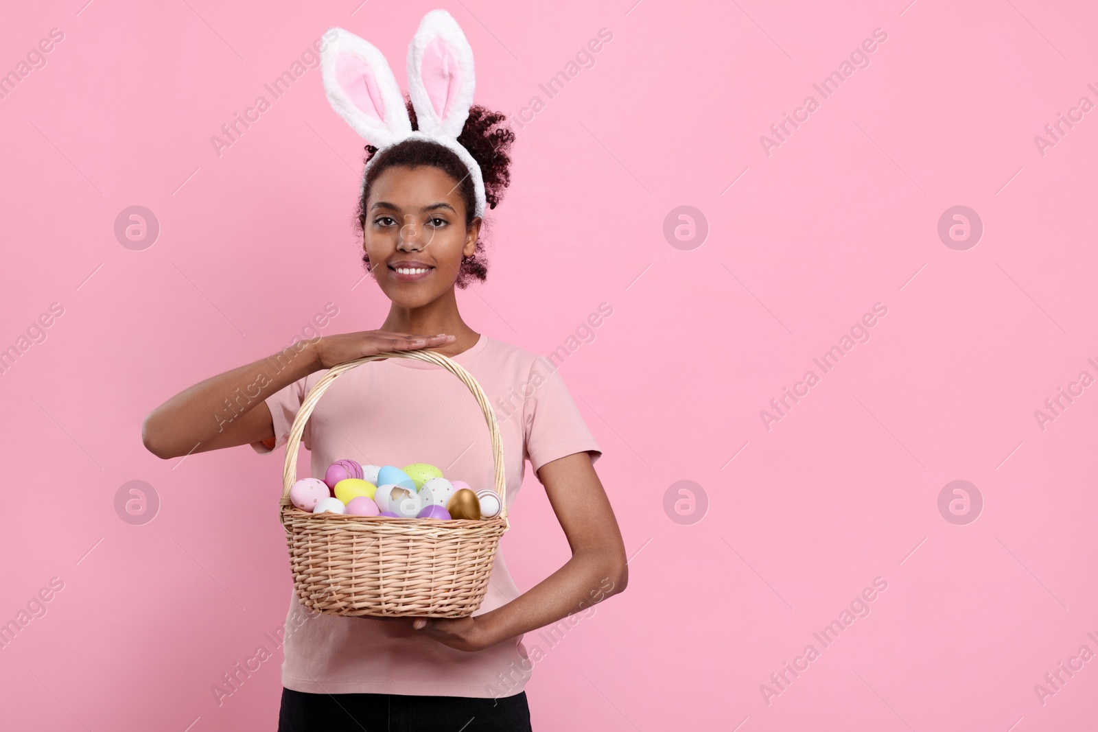 Photo of Happy African American woman in bunny ears headband holding wicker basket with Easter eggs on pink background, space for text