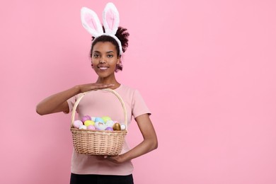 Photo of Happy African American woman in bunny ears headband holding wicker basket with Easter eggs on pink background, space for text