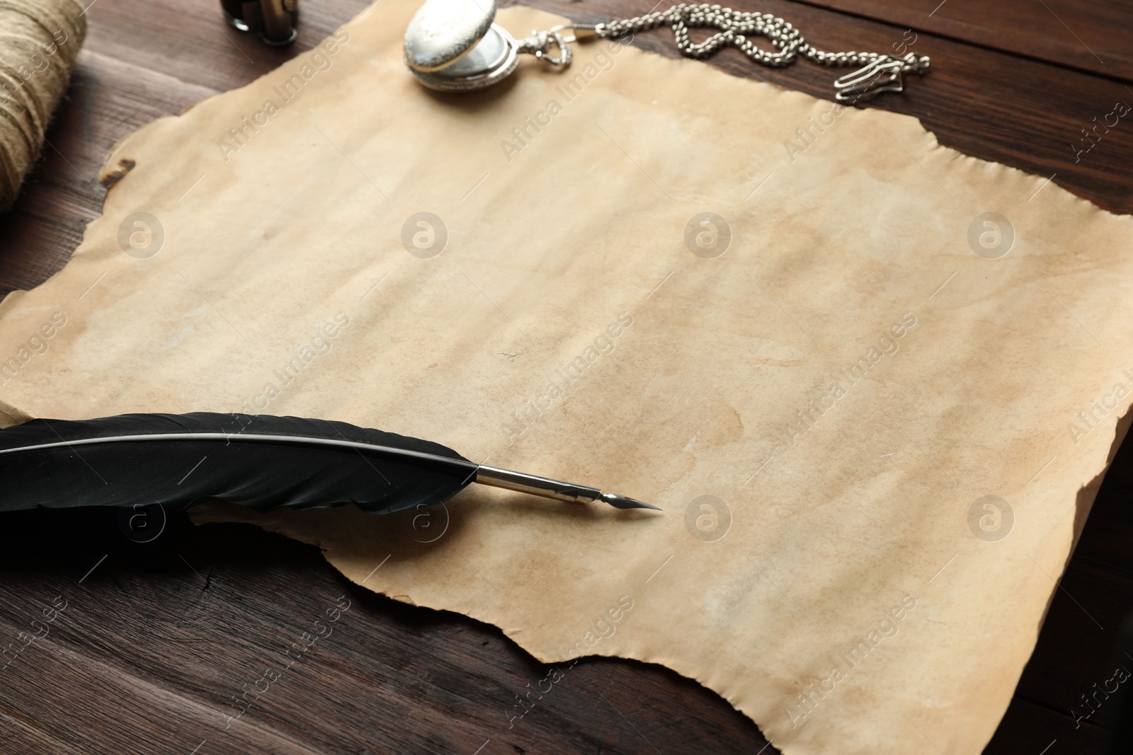 Photo of Sheet of old parchment paper, black feather, inkwell and pocket chain clock on wooden table, closeup
