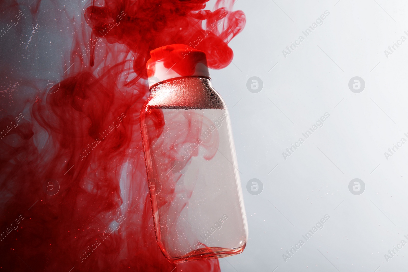 Photo of Bottle of micellar water in liquid with diffusing red paint on light grey background. Space for text