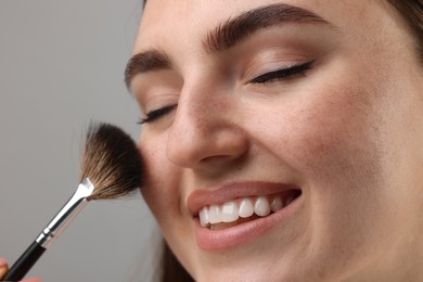 Photo of Smiling woman with freckles applying makeup with brush on grey background, closeup