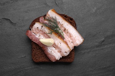 Photo of Tasty pork fatback with rye bread slice, garlic and dill on black table, top view