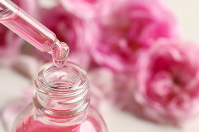 Photo of Dripping rose essential oil into bottle against blurred flowers on table, space for text