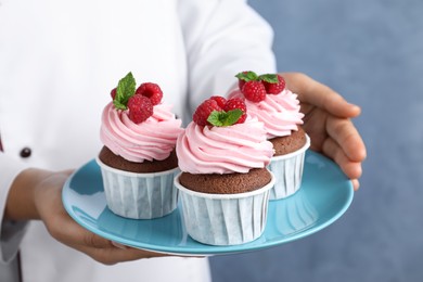 Photo of Pastry chef holding plate with sweet cupcakes on blue background, closeup
