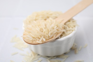 Wooden spoon and bowl with raw rice on white table, closeup