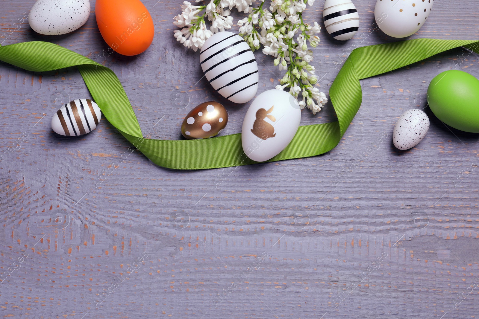 Photo of Many painted Easter eggs, branch with lilac flowers and ribbon on grey wooden table, flat lay. Space for text