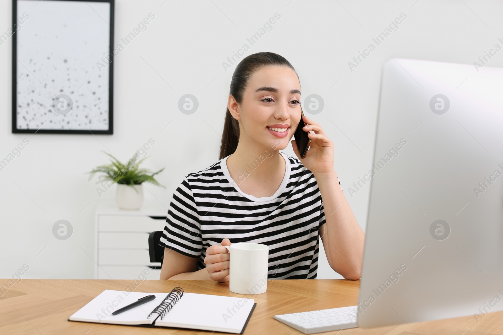 Photo of Home workplace. Happy woman with cup of hot drink talking on smartphone at wooden desk in room