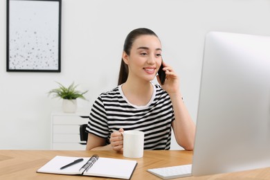 Photo of Home workplace. Happy woman with cup of hot drink talking on smartphone at wooden desk in room