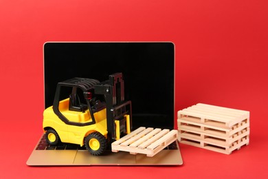 Laptop, toy forklift and wooden pallets on red background