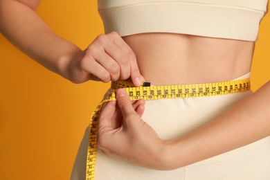 Woman in sportswear measuring waist with tape on yellow background, closeup