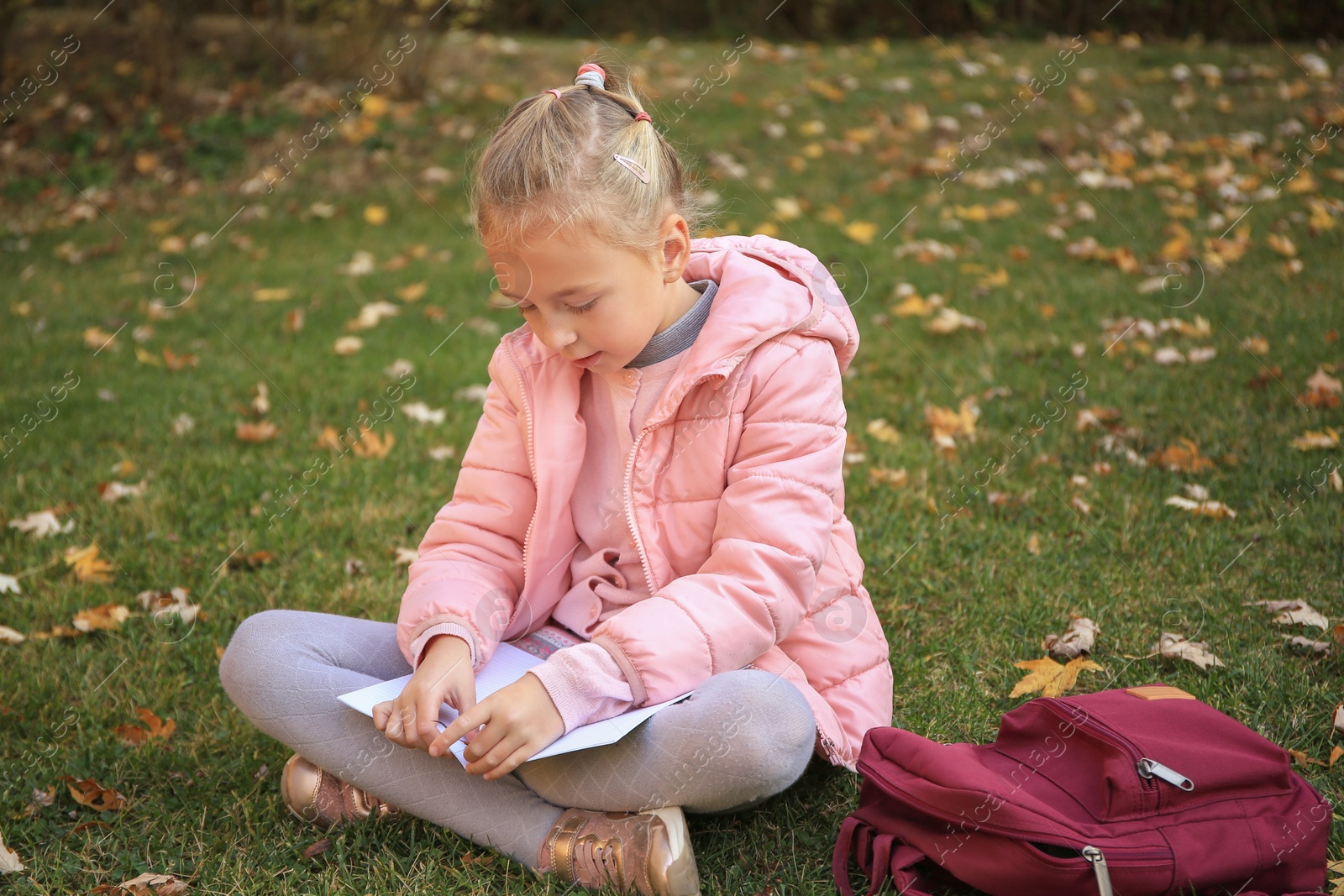 Photo of Cute little girl with copybook and backpack on green grass outdoors