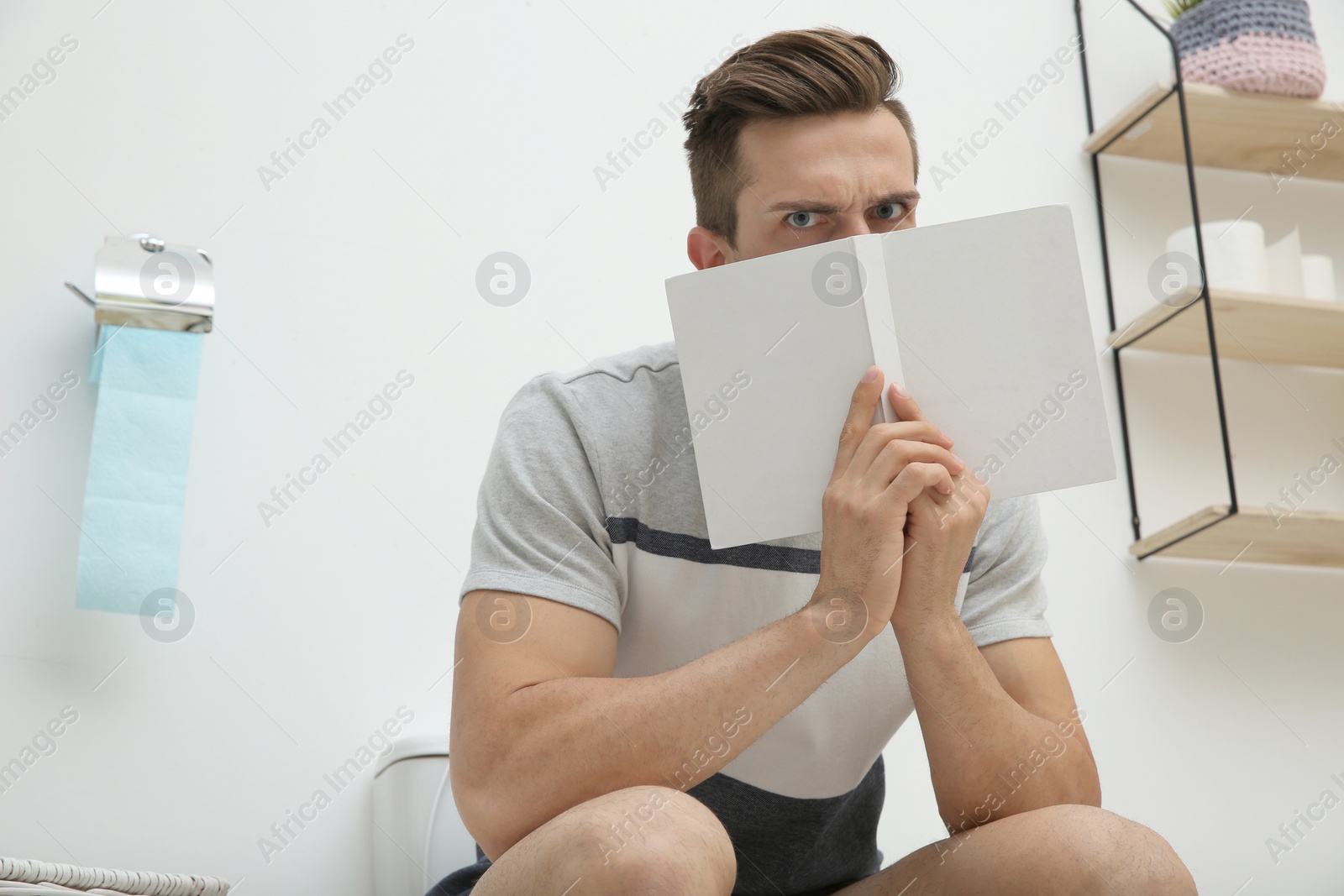 Photo of Young man with book sitting on toilet bowl in bathroom