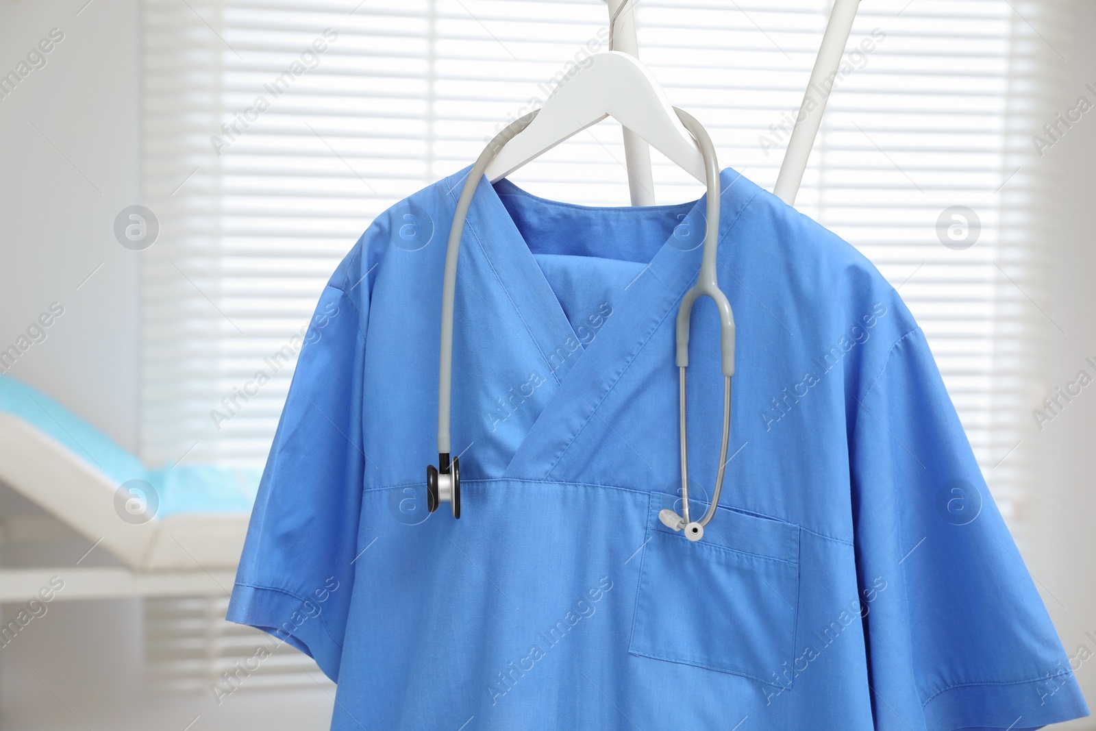 Photo of Blue medical uniform and stethoscope hanging on rack in clinic, closeup