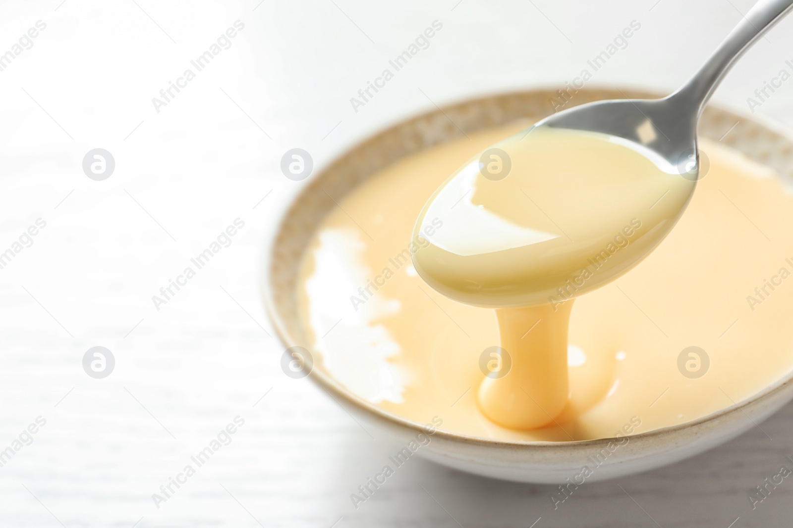 Photo of Spoon of pouring condensed milk over bowl on table, closeup with space for text. Dairy products