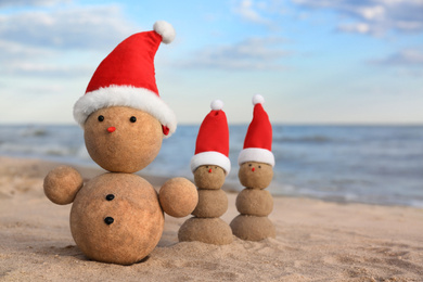 Photo of Snowmen made of sand with Santa hats on beach near sea. Christmas vacation