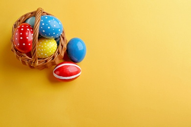 Wicker basket with painted Easter eggs on color background, top view. Space for text