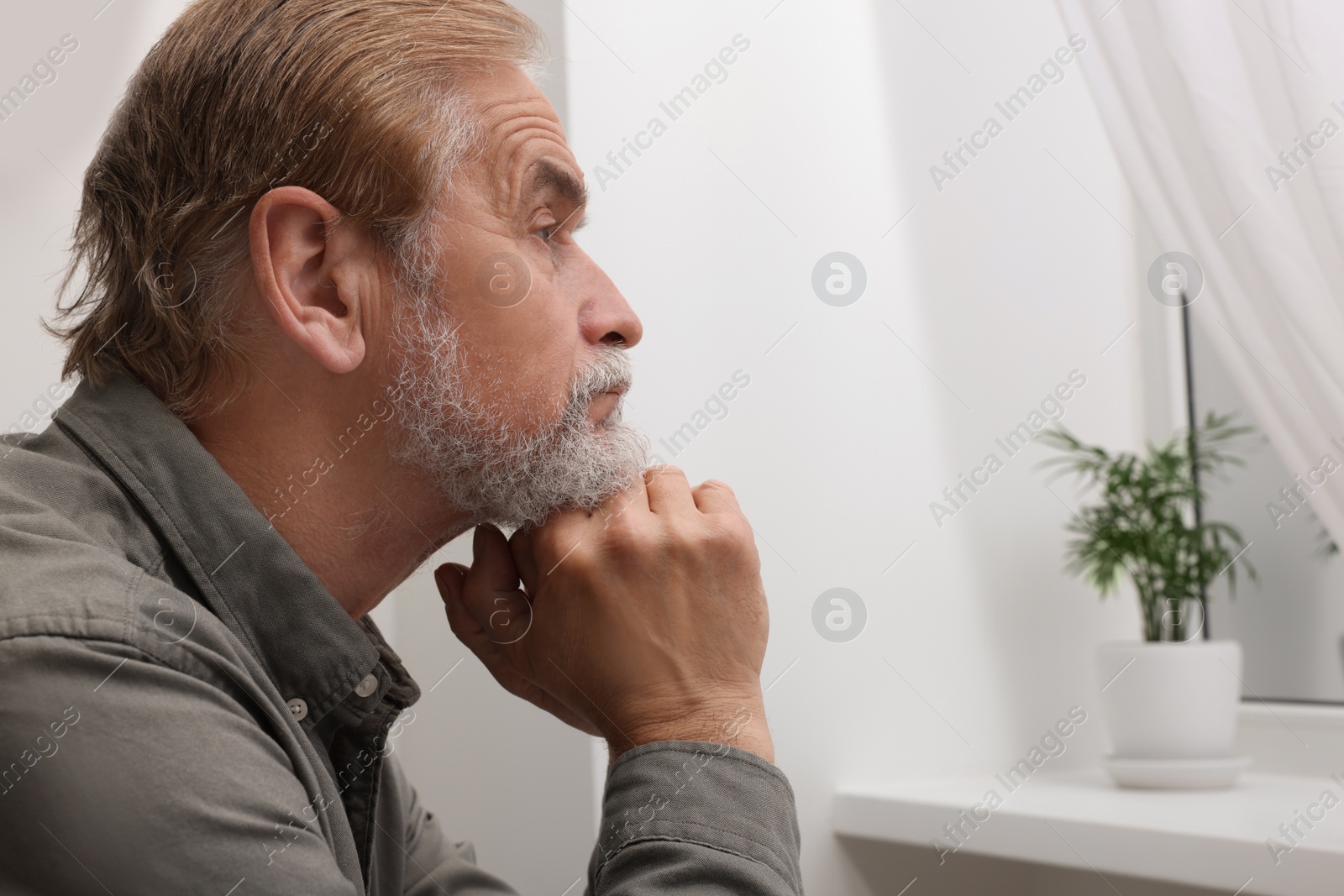 Photo of Upset senior man looking at window indoors, space for text. Loneliness concept