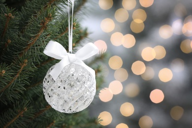 Photo of Shiny white ball with bow hanging on Christmas tree against festive lights, closeup. Space for text