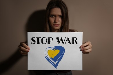 Upset woman holding paper with phrase Stop War near beige wall