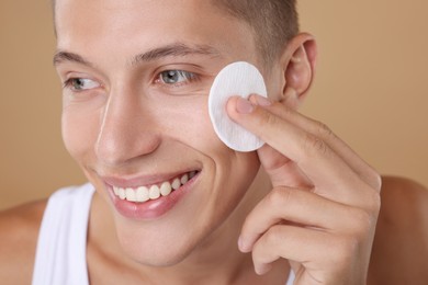 Handsome man cleaning face with cotton pad on beige background, closeup