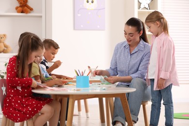 Photo of Nursery teacher and group of cute little children making toys from color paper at desks in kindergarten. Playtime activities
