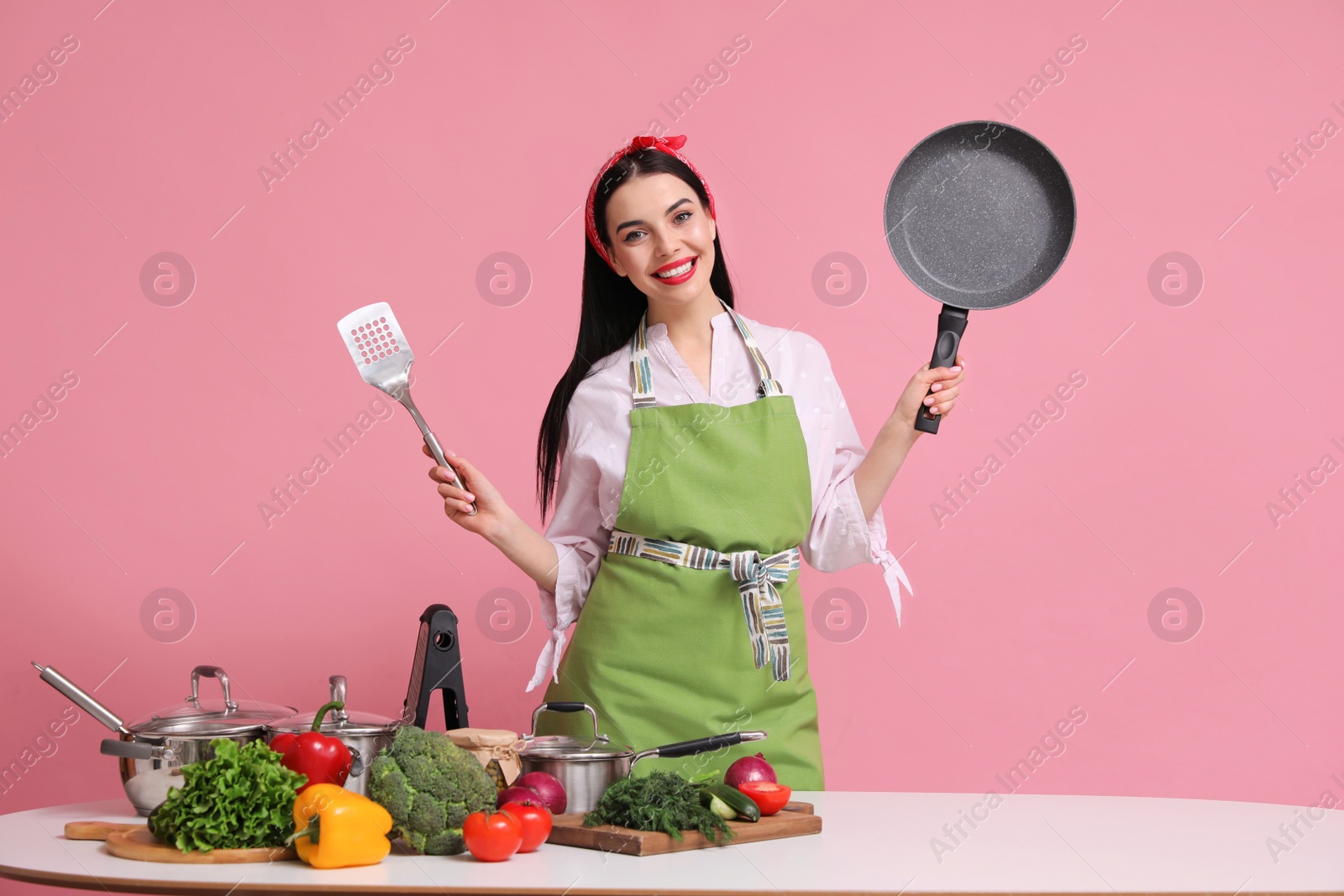 Photo of Young housewife with pan, spatula and products on pink background