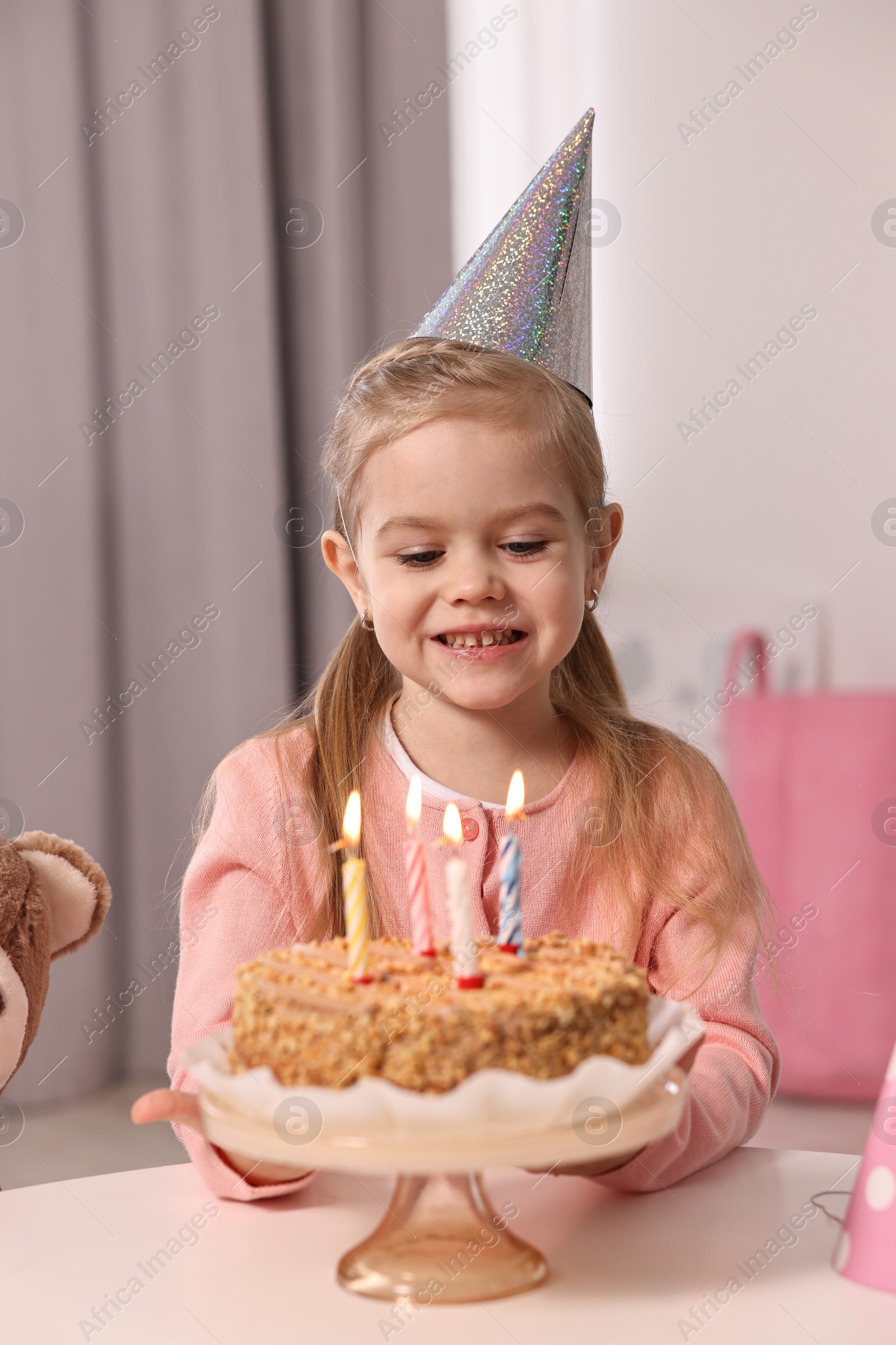 Photo of Cute girl in party hat with birthday cake at table indoors