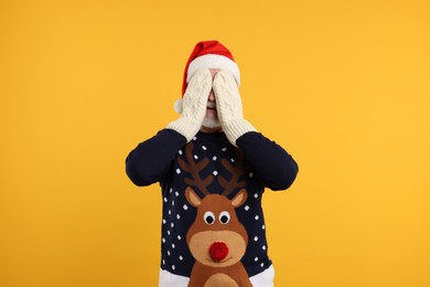Photo of Senior man in Christmas sweater and Santa hat covering face with hands in knitted mittens on orange background