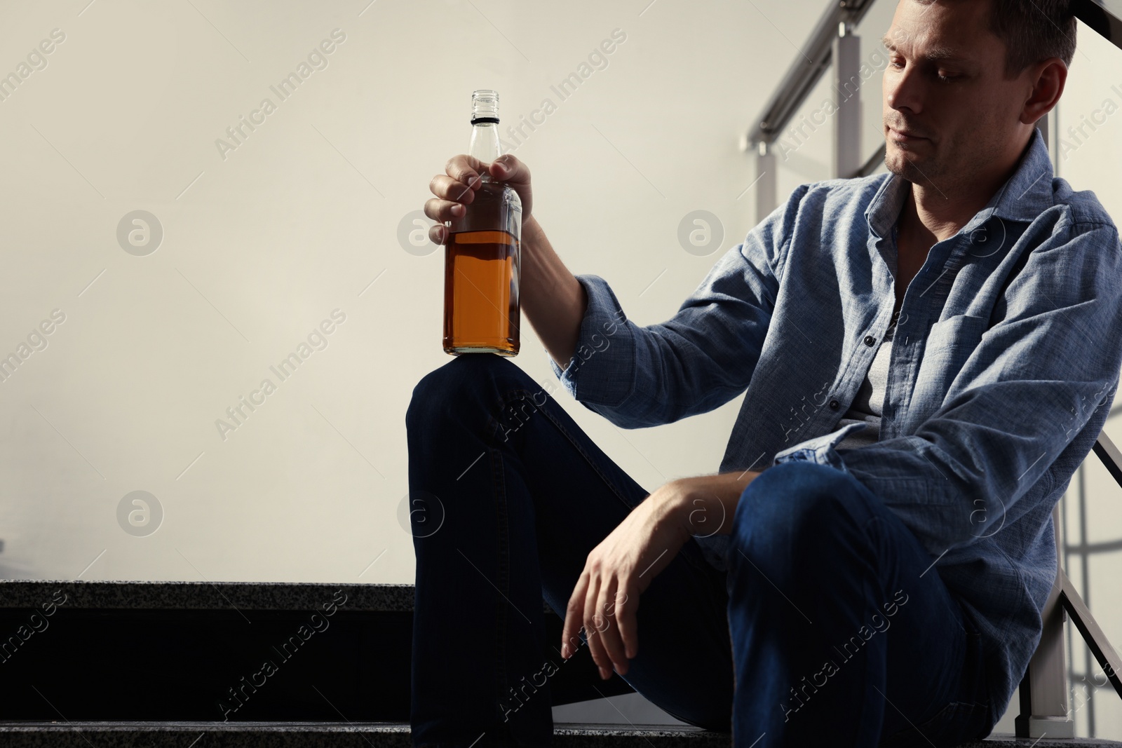 Photo of Addicted man with alcoholic drink on stairs indoors. Space for text