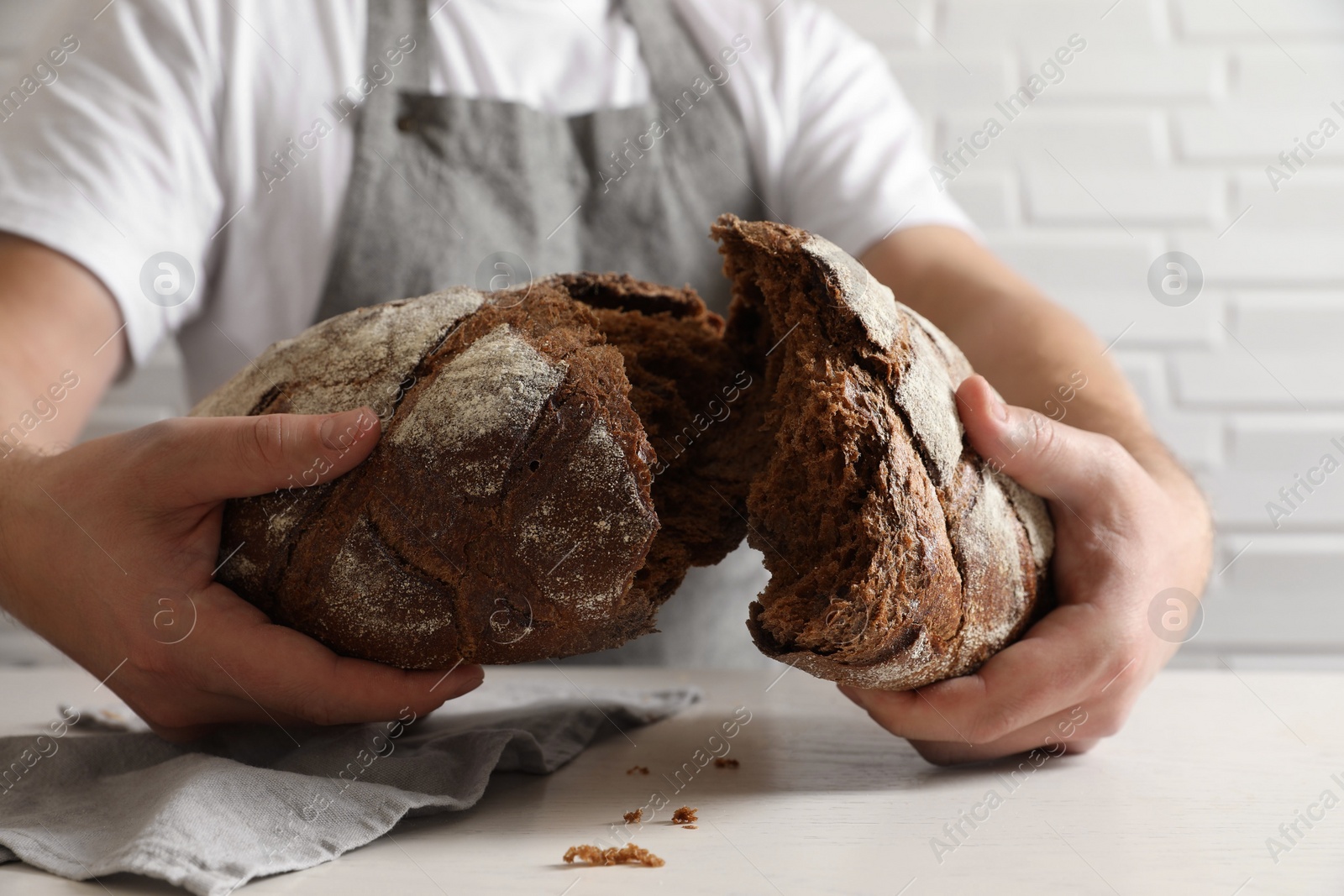 Photo of Man breaking loaf of fresh bread at white table near brick wall, closeup