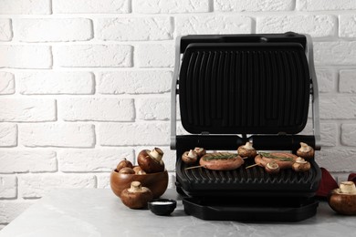 Photo of Electric grill with homemade sausages, rosemary and mushrooms on marble table. Space for text