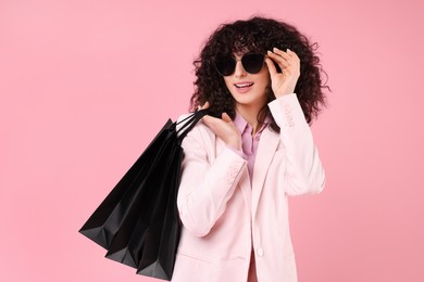 Happy young woman with shopping bags and stylish sunglasses on pink background