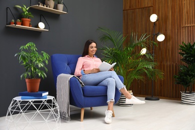Photo of Happy woman reading book in armchair at home. Indoor plants for trendy interior design