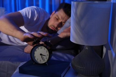 Photo of Sleepy man turning off alarm clock in dark room. Bedtime