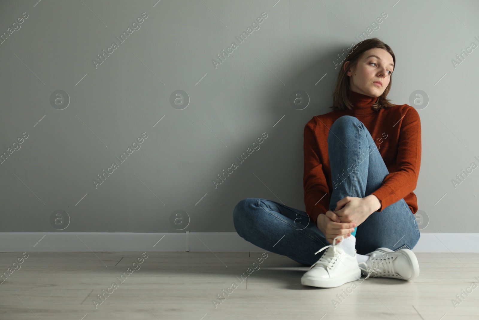 Photo of Sad young woman sitting on floor near grey wall indoors, space for text