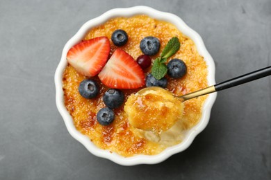 Photo of Taking delicious creme brulee with berries and mint from bowl at grey table, top view